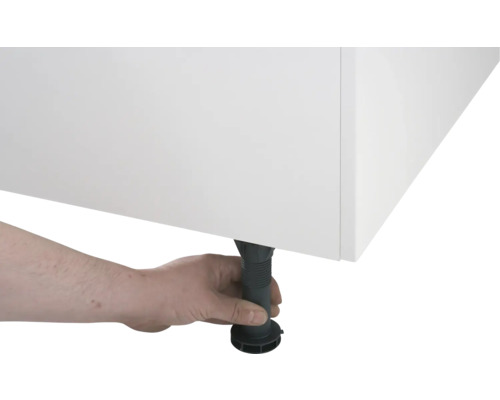 Une main ajuste un pied de meuble noir réglable en hauteur sous un meuble blanc.