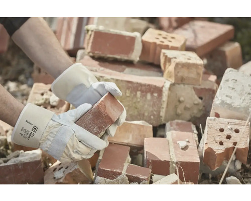 Eine Person mit Arbeitshandschuhen hält einen Ziegelstein vor einem Haufen Ziegelsteine.