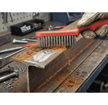 Une main gantée nettoie un profilé en acier rouillé avec une brosse métallique.