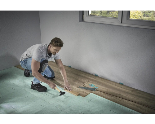 Un homme pose du stratifié avec un marteau et une cale de frappe sur une isolation phonique dans une pièce.