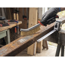 Eine Person benutzt eine Drahtbürste zur Rostentfernung auf einem Metallstück neben einem Rollbandmaß.