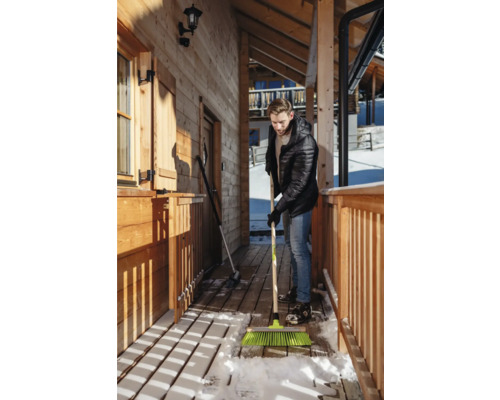 Mann kehrt Schnee mit einem Besen vor einem Holzhaus