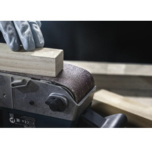 Gros plan d'une ponceuse à bande ponçant une pièce de bois, manipulée par une personne gantée.