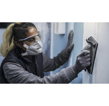 Eine Person trägt Schutzausrüstung und verwendet ein Schleifbrett zum Schleifen einer Wand.