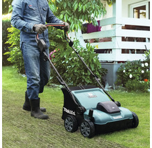 Une personne avec des gants et des bottes pousse un scarificateur Makita sur une pelouse dans un jardin lumineux et entretenu.