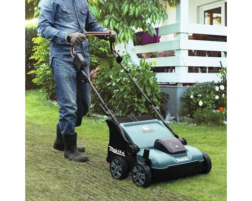 Une personne avec des gants et des bottes pousse un scarificateur Makita sur une pelouse dans un jardin lumineux et entretenu.