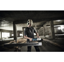 Handwerker mit Schutzhelm und Gehörschutz bearbeitet einen Metallträger mit einem Makita Winkelschleifer in einem Rohbau aus Beton.