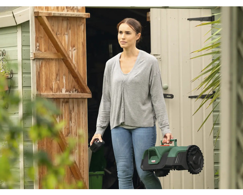 Femme portant un robot tondeuse devant une remise de jardin.