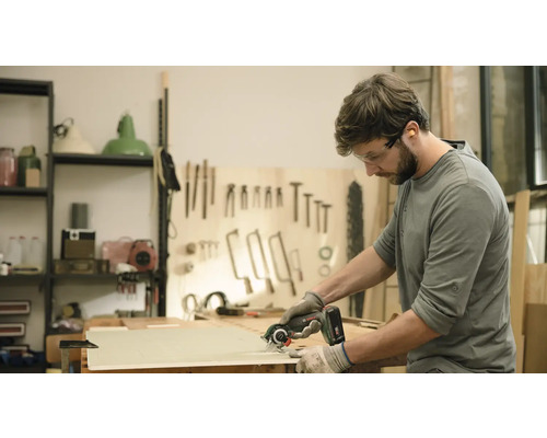 Ein Mann schneidet mit einer Handkreissäge ein Holzbrett. Er trägt dabei eine Schutzbrille und Handschuhe.