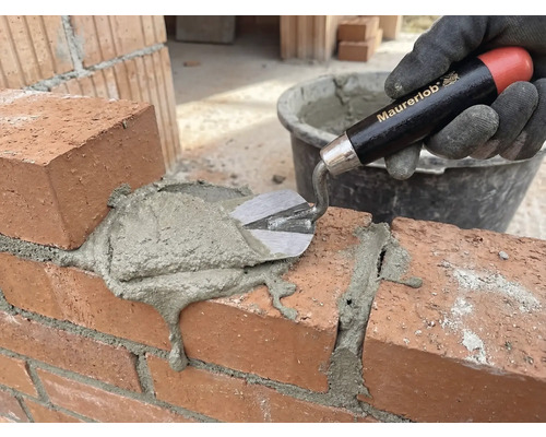 Eine Hand mit Handschuh trägt mit einer Maurerkelle Mörtel auf eine Ziegelmauer auf