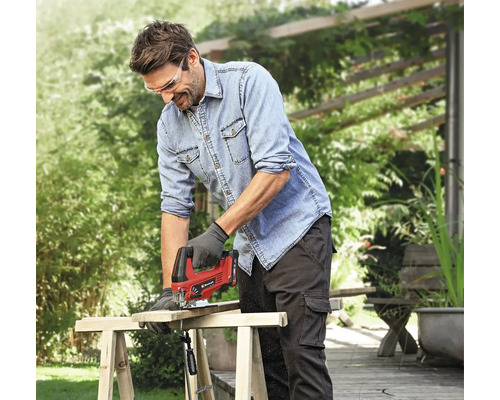Un homme scie une planche de bois à l'extérieur avec une scie sauteuse. Il porte des lunettes de protection, une blouse de travail et des gants de travail.