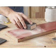 Une main ponce une planche de bois laquée avec un non-tissé abrasif sur un bloc à poncer. De la poussière est visible sur le bois.