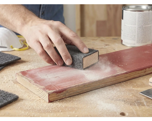 Une main ponce une planche de bois laquée avec un non-tissé abrasif sur un bloc à poncer. De la poussière est visible sur le bois.