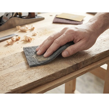 Main traitant une planche de bois avec un tampon abrasif sur un établi lumineux avec un rabot et des copeaux.