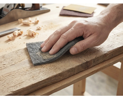 Main traitant une planche de bois avec un tampon abrasif sur un établi lumineux avec un rabot et des copeaux.