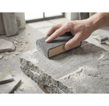 Une main guide un bloc de ponçage en bois avec une toison abrasive grise sur la surface d'une pierre naturelle brute.