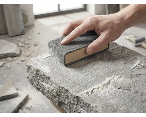 Une main guide un bloc de ponçage en bois avec une toison abrasive grise sur la surface d'une pierre naturelle brute.