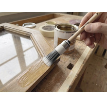 Anwendung einer Holzlasur auf einem Holzrahmen mit einem Rautner Rundpinsel auf einer hellen Werkbank mit Farbdose und Klebeband.