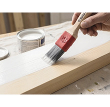 Un pinceau avec manche en bois et virole rouge applique de la peinture blanche sur une poutre en bois. Un pot de peinture est visible en arrière-plan.