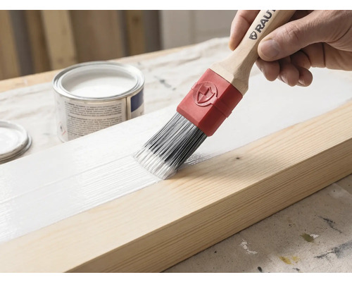 Un pinceau avec manche en bois et virole rouge applique de la peinture blanche sur une poutre en bois. Un pot de peinture est visible en arrière-plan.