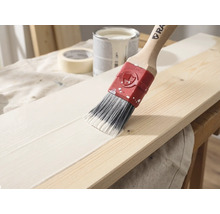 Un pinceau avec un manche en bois applique de la peinture blanche sur une planche en bois, un pot de peinture et du ruban adhésif en arrière-plan.