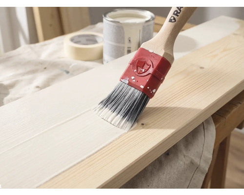 Un pinceau avec un manche en bois applique de la peinture blanche sur une planche en bois, un pot de peinture et du ruban adhésif en arrière-plan.