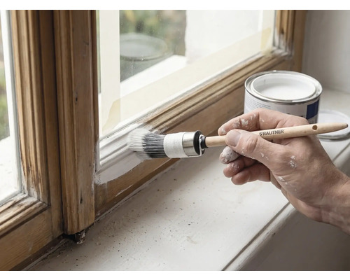 Hand streicht einen Holzfensterrahmen weiß mit einem Rautner Ringpinsel, neben einer geöffneten Farbdose.