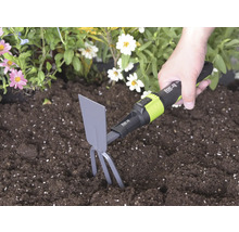 Main utilisant une serfouette for_q avec dents et lame dans une terre sombre à côté de fleurs pour ameublir le sol.