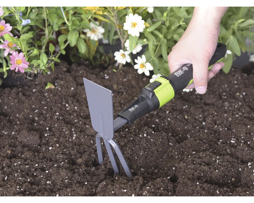 Main utilisant une serfouette for_q avec dents et lame dans une terre sombre à côté de fleurs pour ameublir le sol.