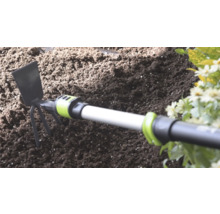 Serfouette avec lame en métal et trois dents dans du terreau sombre à côté de fleurs.