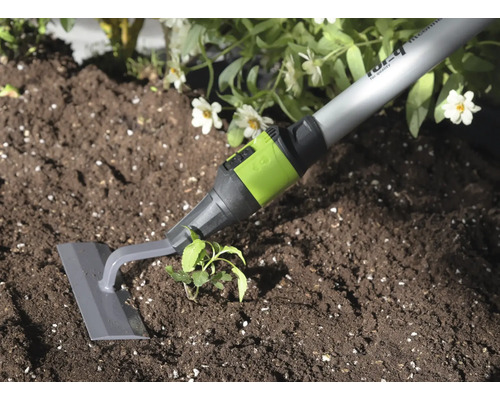 Sarcloir de jardin avec lame grise et raccord vert utilisé pour désherber la terre à côté d'une plante.