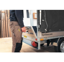Person in Arbeitskleidung öffnet die Heckklappe eines Humbaur Pkw-Anhängers in einer hellen Werkstatt. Humbaur Logo.