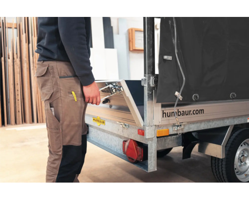Person in Arbeitskleidung öffnet die Heckklappe eines Humbaur Pkw-Anhängers in einer hellen Werkstatt. Humbaur Logo.