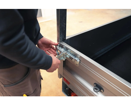 Person bedient einen Winkelhebelverschluss an einer silbernen Pkw-Anhänger-Bordwand aus Metall.