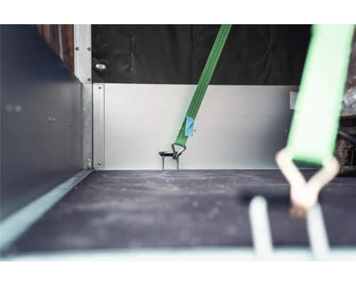 Grüner Spanngurt mit Haken in einer Verzurröse auf der Ladefläche eines Anhängers zur Ladungssicherung.