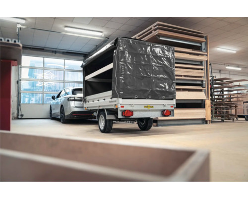 Einachsiger Humbaur PKW-Anhänger mit grauer Hochplane in einer Lagerhalle neben Holzregalen und einem Auto.