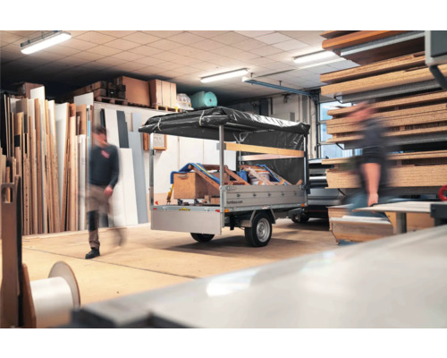 Humbaur Anhänger mit schwarzer Hochplane in einer hellen, gut organisierten Werkstatt mit Holzplatten und Baumaterialien.
