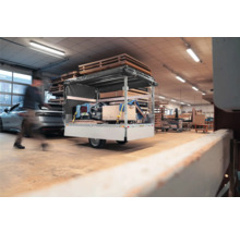 Einachsiger Pkw-Anhänger mit Hochplane und Ladung in einer hellen Werkstatt neben Holzstapeln und einem Auto.