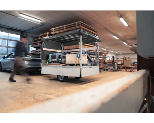 Einachsiger Pkw-Anhänger mit Hochplane und Ladung in einer hellen Werkstatt neben Holzstapeln und einem Auto.
