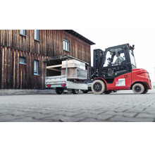 Ein roter Gabelstapler belädt einen silbernen Pkw-Anhänger mit einer Palette vor einem hölzernen Gebäude.