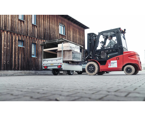 Ein roter Gabelstapler belädt einen silbernen Pkw-Anhänger mit einer Palette vor einem hölzernen Gebäude.