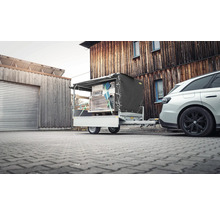 Pkw-Anhänger beladen mit verzurrten Waren und grauer Plane, angekoppelt an ein Auto vor einem Holzgebäude auf gepflastertem Boden.