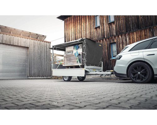 Pkw-Anhänger beladen mit verzurrten Waren und grauer Plane, angekoppelt an ein Auto vor einem Holzgebäude auf gepflastertem Boden.