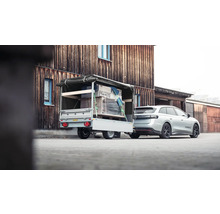 Auto mit beladenem Anhänger, Ladung gesichert mit Spanngurten und Plane, vor einem Holzgebäude.