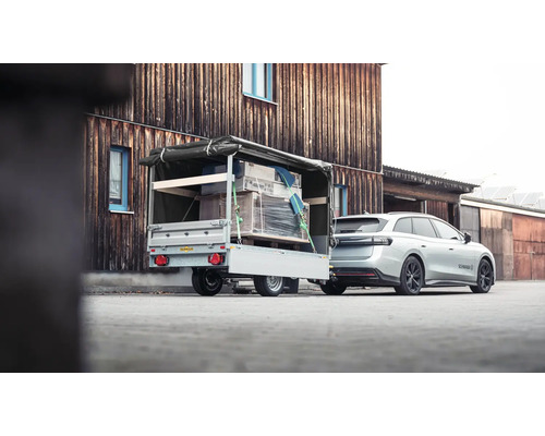 Auto mit beladenem Anhänger, Ladung gesichert mit Spanngurten und Plane, vor einem Holzgebäude.