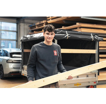 Ein Mann hält einen langen Holzbalken vor einem Anhänger in einem gut sortierten Holzlager.