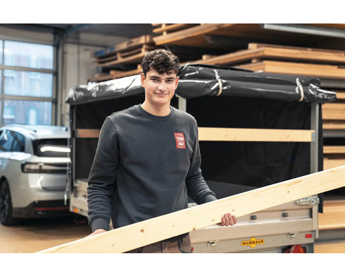 Ein Mann hält einen langen Holzbalken vor einem Anhänger in einem gut sortierten Holzlager.