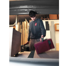 Ein Handwerker in einer Werkstatt trägt zwei Werkzeugkoffer aus Kunststoff und platziert einen davon auf einer Arbeitsfläche vor Holzplatten.