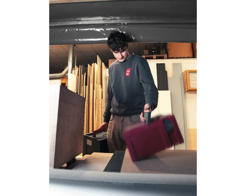 Ein Handwerker in einer Werkstatt trägt zwei Werkzeugkoffer aus Kunststoff und platziert einen davon auf einer Arbeitsfläche vor Holzplatten.