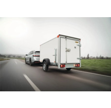 Une remorque fourgon Humbaur blanche avec des portes arrière est tractée par une voiture sur une route de campagne.